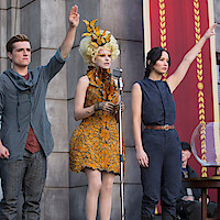 Three people stand on a stage in front of a microphone and banners, two with arms raised in a salute; urban setting, one in a decorative yellow dress, the others in simple outfits, evoking a ceremonial or public event atmosphere.