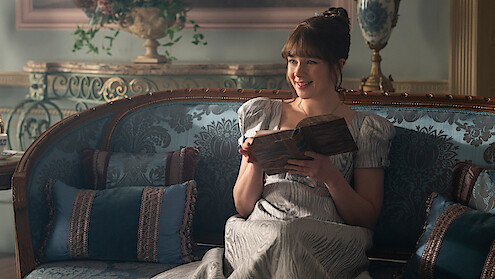 Woman in historical dress sitting on ornate blue couch in an elegant, vintage room, smiling while reading a book, surrounded by decorative cushions and classical furnishings.