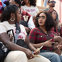Two young women talk in a stadium crowd, one holding cash, both wearing college apparel. Other spectators in casual outfits and sunglasses sit on tiered steps, enjoying an outdoor event under bright daylight.