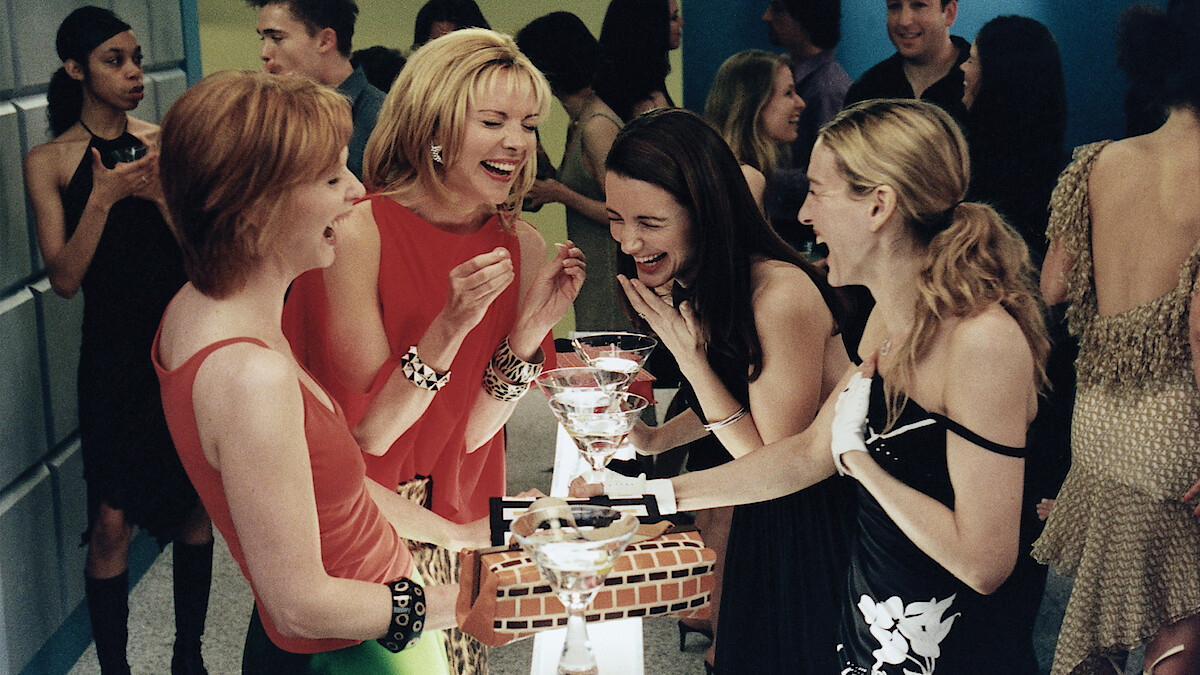 Four women dressed stylishly laugh and enjoy cocktails together around a small table at a lively indoor party with people socializing in the background.