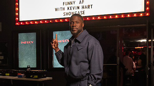 Kevin Hart in a blue jacket stands inside a comedy club, pointing upward at a lit marquee sign that reads “Funny AF with Kevin Hart Showcase,” with tables, equipment, and an audience in the background.