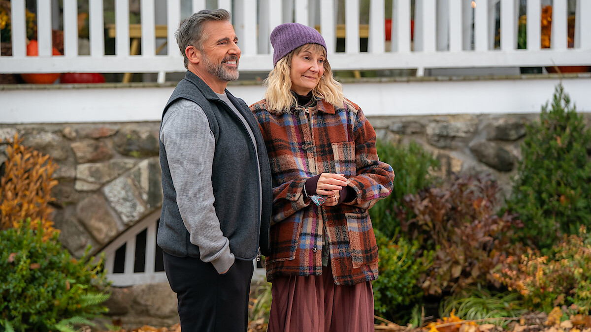 A man and woman stand outdoors in autumn clothing, smiling in front of a stone wall and white railing, surrounded by fall foliage and plants.