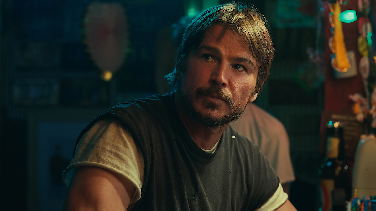 Man with short brown hair and beard sits at a dimly-lit bar, wearing a dark T-shirt over a light shirt. Bar items and ambient lighting are visible in the colorful, casual setting.