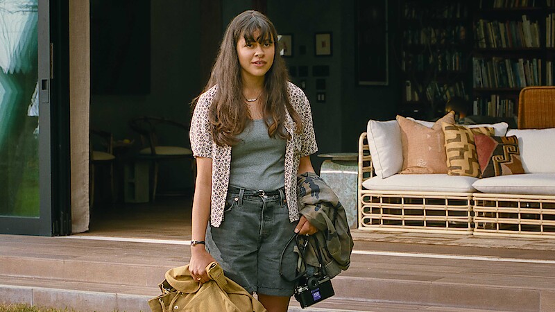 Young woman with long hair stands outside a cozy, modern house holding a coat, bag, and camera. The relaxed setting features a wicker sofa and bookshelves, evoking a calm, casual, and welcoming atmosphere.