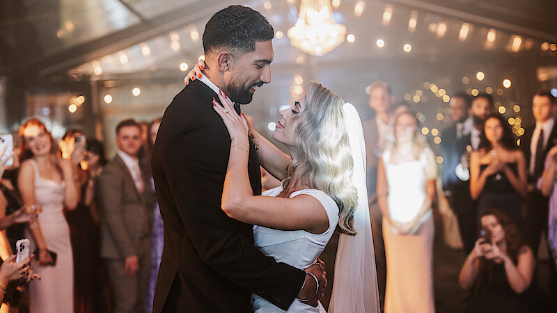 Bride and groom share a first dance at their wedding, surrounded by guests in formal attire under string lights inside a decorated venue.