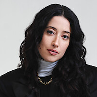 Taylor Ortega photographed in front of a white backdrop wearing a black jacket with strong shoulders, white turtle neck layered under a black shirt, and a gold chain with her dark curly hair cascading around her face.