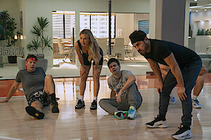 Four tired people in sportswear resting on an indoor basketball court, with office spaces and large windows in the background, suggesting a recreational break in a modern office environment.
