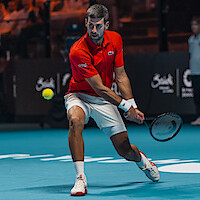 Male tennis player in a red shirt and white shorts hitting a backhand shot on an indoor hard court during a match, with the tennis ball in motion and an audience in the background.