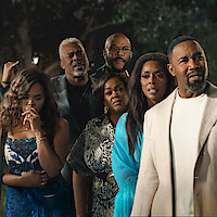 A group of six adults dressed in formal and semi-formal attire stand close together outdoors at night, looking intensely ahead with concerned or serious expressions, trees and dark background behind them.
