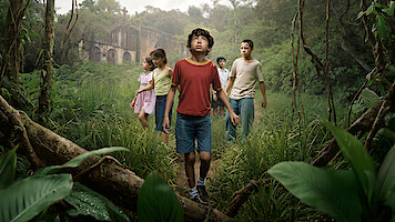 A group of children walk through dense green jungle foliage, looking around curiously. In the background, a large, old, abandoned stone building stands partially hidden by mist and trees, creating an adventurous and mysterious atmosphere.