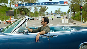 Man driving a classic blue convertible near Dodger Stadium entrance on a sunny day, with a relaxed vibe, palm trees and stadium signs visible, scene feels casual and laid-back.