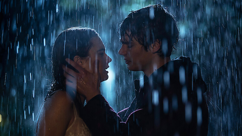 Two people stand close together in the rain at night, gazing into each other's eyes as one gently holds the other’s face, surrounded by falling water and dramatic lighting, creating a romantic and emotional atmosphere.