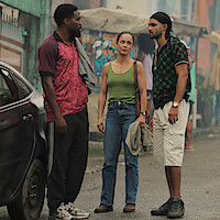 Three people stand talking on a street near a parked car, with colorful buildings and market stalls in the background, dressed casually in a lively urban outdoor setting.