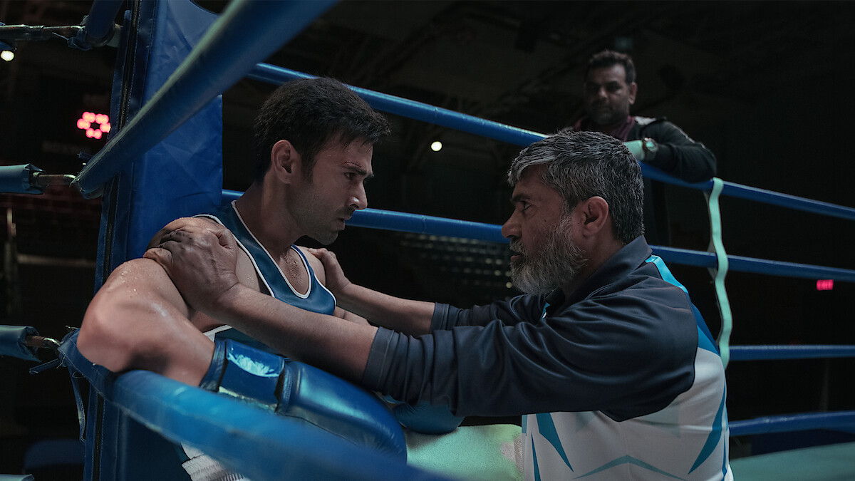 A boxer sits in a boxing ring corner, wearing blue and white gear, while his coach kneels in front of him giving advice; another person stands in the background, dimly lit arena.