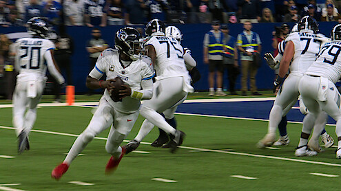 Football quarterback in white uniform running with the ball on a green field during a game, with teammates blocking and stadium spectators in the background.
