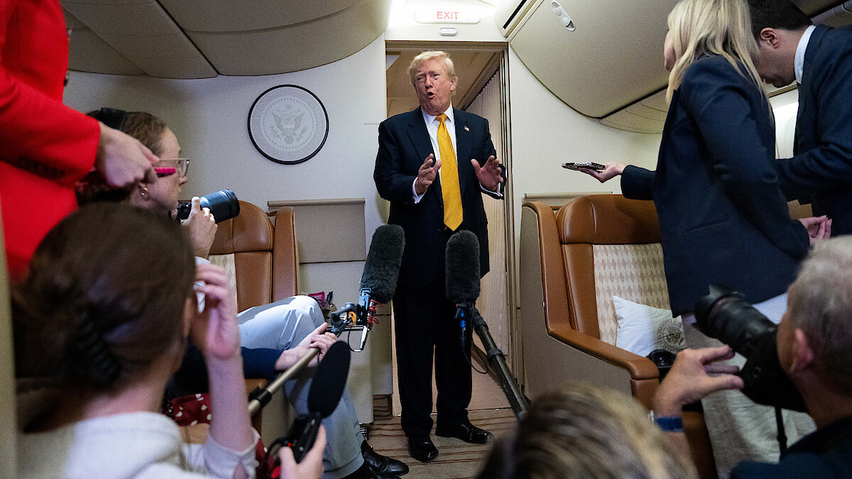 Man in suit speaks to reporters holding microphones and recording devices inside an airplane cabin, with leather seats and presidential seal visible in the background.