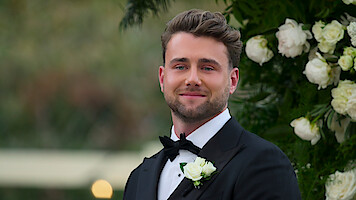 A man in a black tuxedo with a white boutonniere smiles outdoors near an arrangement of white flowers and greenery, suggesting a formal or wedding setting.