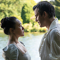 A man and woman in period clothing stand closely facing each other near a lake, surrounded by greenery and trees on a bright day, suggesting a romantic or dramatic moment outdoors.