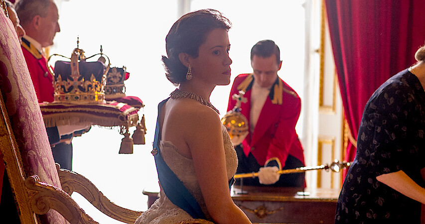 Claire Foy as the Queen in The Crown. She's sitting behind a desk with the royal crown behind her