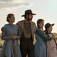 A family of four in prairie-style clothing stands close together in a grassy field at sunset, looking toward the horizon with concerned expressions, evoking a historical or frontier setting.