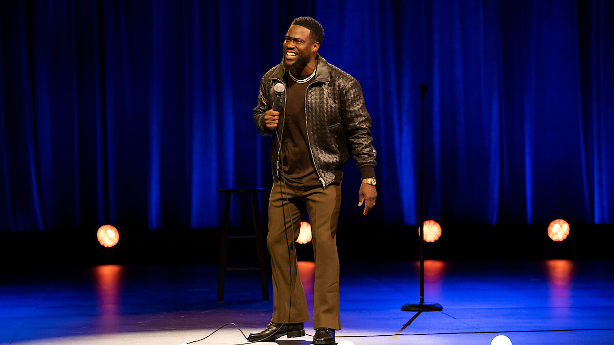Comedian performing stand-up on stage with blue curtains, holding a microphone and smiling. Stage lights and a stool are visible in the background.