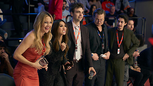 Five people standing and cheering at an indoor event, wearing semi-formal clothes with ID badges, in a crowded arena with audience members visible in the background.