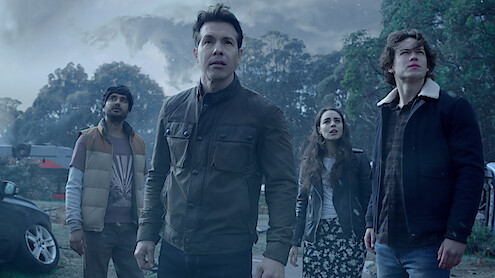 Four people standing outdoors in a tense atmosphere, surrounded by cars and trees under a stormy, cloudy sky, all looking up with concerned expressions.