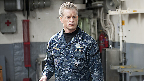 A man in a blue digital camouflage Navy uniform stands indoors on a ship, surrounded by industrial equipment, pipes, and emergency gear, looking serious and focused.