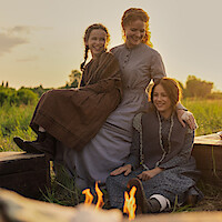 Three women in vintage pioneer dresses sit together outside on wooden benches by a campfire, smiling and enjoying a sunset in a grassy field.