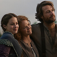 Three people dressed in period clothing standing outdoors, smiling and embracing, with soft natural light and a blurred background suggesting a rural or countryside setting.