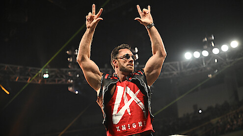 A muscular man wearing sunglasses and a red vest stands in an arena with bright lights, raising both arms and making hand gestures, with an audience and stage lighting visible in the background.