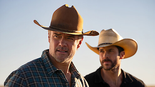 Two men wearing cowboy hats stand outdoors under a clear sky, one in a plaid shirt in the foreground, the other blurred in the background, giving a sense of a rural or Western environment.