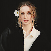 Woman with wavy blonde hair and a white flower tucked behind her ear, wearing a black coat over a cream shirt, poses in front of a dark background with a soft, natural expression.