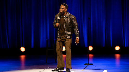 Comedian performing stand-up on stage with blue curtains, holding a microphone and smiling. Stage lights and a stool are visible in the background.