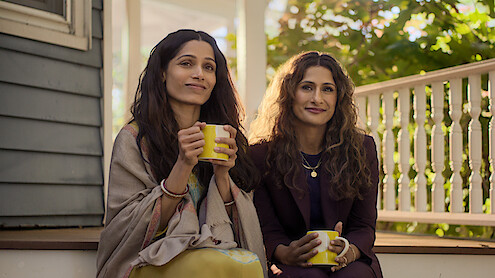 Two women sit together on a sunlit porch, holding yellow mugs and smiling. Shrubs and a white railing are visible in the background, creating a relaxed, inviting environment.
