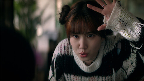 Young woman indoors with plants in the background, wearing a black and white sweater with frayed sleeves, hair in two buns, raising her hand and making a playful expression, as if acting or interacting animatedly.