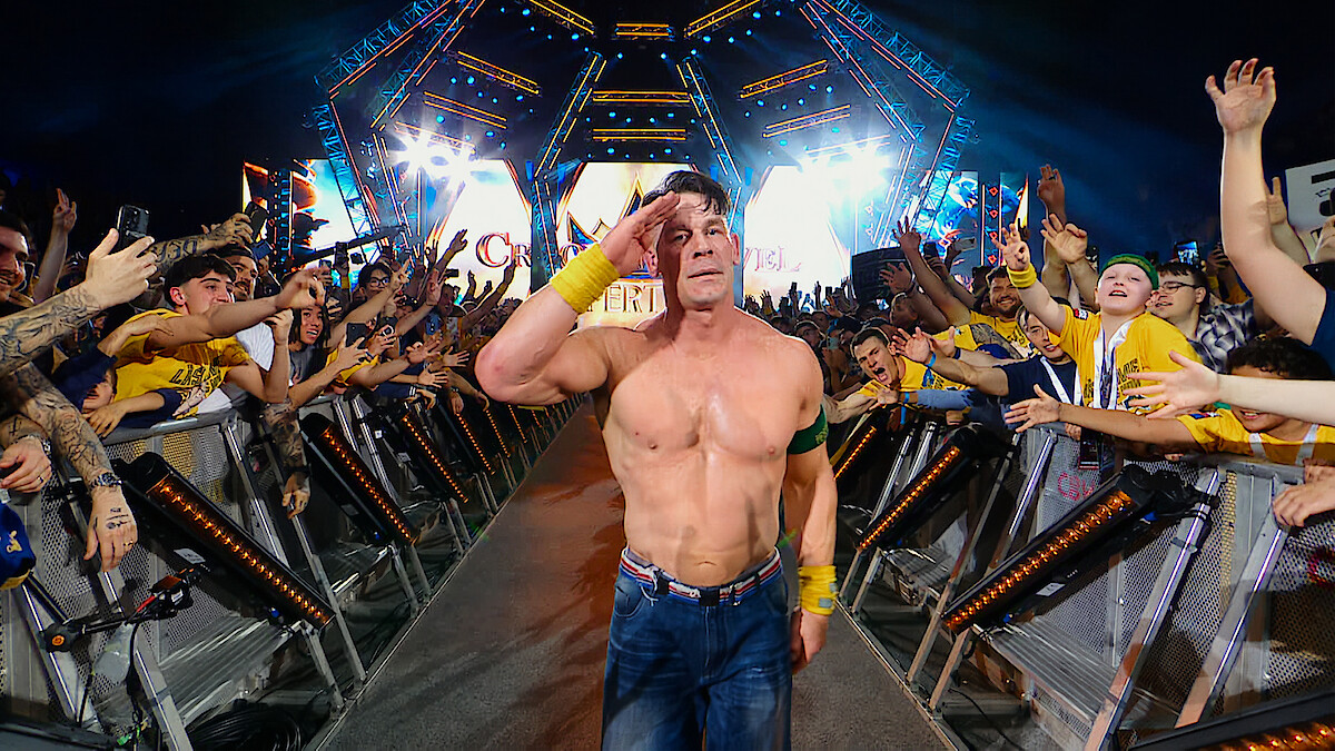 Shirtless wrestler saluting and walking down a ramp, surrounded by cheering fans at a brightly lit arena event, with colorful lights and stage setup in the background.
