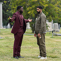 Two men talk in a cemetery; one wears a burgundy suit with arms open in a questioning gesture, while the other in casual clothes holds a coffee cup. Tombstones and trees are visible in the background.