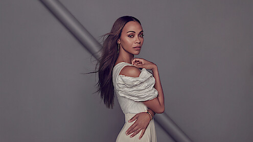 Zoe Saldaña stands against a gray background wearing an ensemble made of different shades of cream along with some gold jewelry, facing the camera with her hand touching her shoulder. 