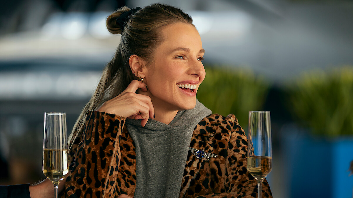 Smiling woman in leopard print coat sitting at a table with champagne glasses in a bright, modern setting, plants blurred in the background, appearing relaxed and joyful.