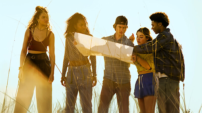 Five young people stand in tall grass outdoors, closely examining a large map together in bright daylight, appearing focused and collaborative in a natural setting.