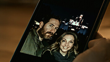 A man and woman smiling in a selfie photo on a smartphone, taken at night in front of an illuminated castle, likely at an amusement park. Hand holding phone visible in foreground.