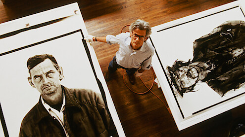An artist stands between two large black-and-white portraits on tables in a studio with wooden floors, looking up at the camera; the environment is bright and spacious, emphasizing the creative setting and detailed artwork.