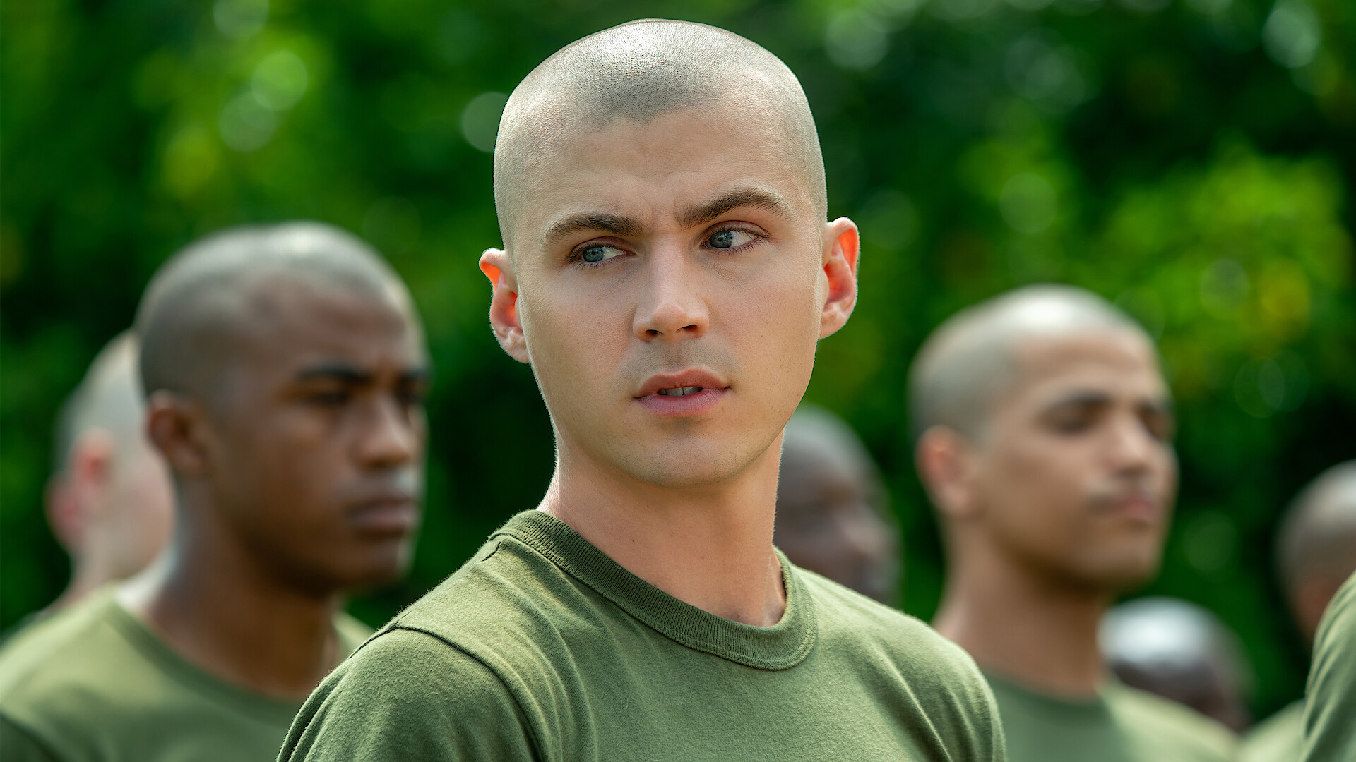 Young man with shaved head in green military shirt stands outdoors among other recruits with similar haircuts, blurred green foliage in background, serious expressions, appearing to be in a training or boot camp setting.