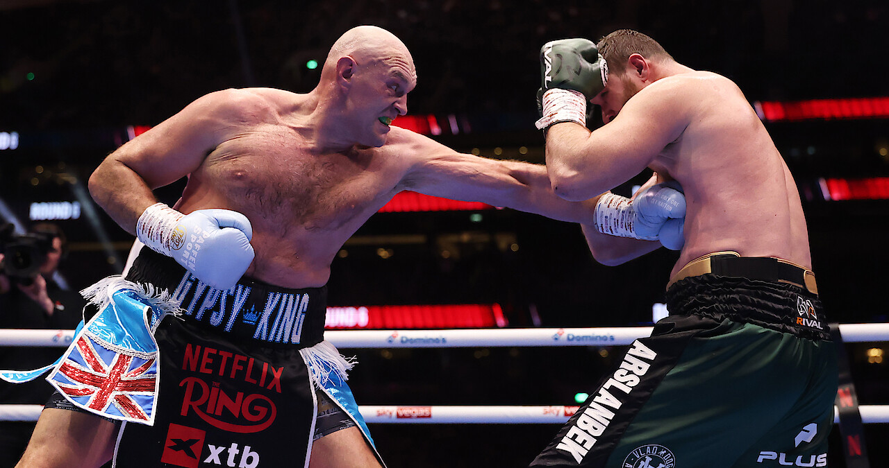 Tyson Fury and Arslanbek Makhmudov in a brightly lit boxing ring, one delivering a punch while the other blocks with his arms up, surrounded by a cheering audience in a large arena.