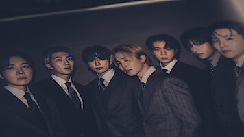 Seven young men wearing formal suits pose together in a dimly lit, dark environment, standing closely for a group portrait with serious and calm expressions.
