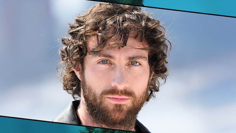 A man with curly brown hair and a beard is outdoors, wearing a black jacket, with a soft blurred background of blue sky and possibly distant mountains.