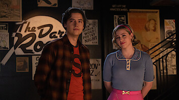 Cole Sprouse as Jughead Jones and Lili Reinhart as Betty Cooper stand together smiling in Season 7 of 'Riverdale.'