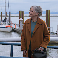 Sally Field as Tova in a brown coat standing by a railing at a marina, with sailboats docked and calm water in the background under a cloudy sky.