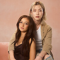 Two people pose together in a studio with a soft pink background; one sits on a pink couch wearing glasses and a dark shirt, while the other, with long blonde hair and a tan jacket, sits close behind, both looking at the camera.